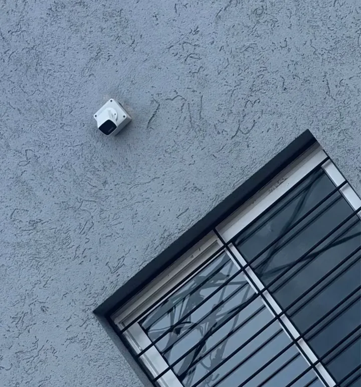 Cámara de seguridad exterior vista desde abajo, montada sobre la fachada junto a una ventana con rejas