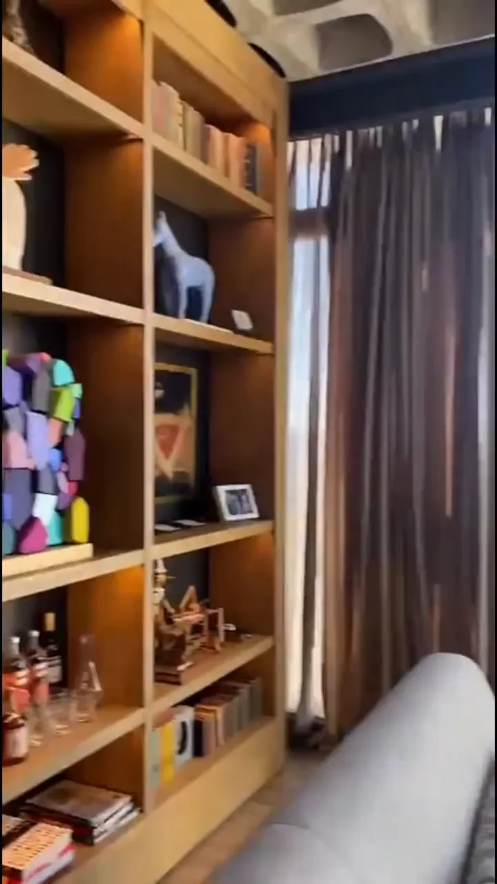 Biblioteca empotrada con objetos de colección, luz cálida embutida en cada estante y ventanal con cortina de fondo