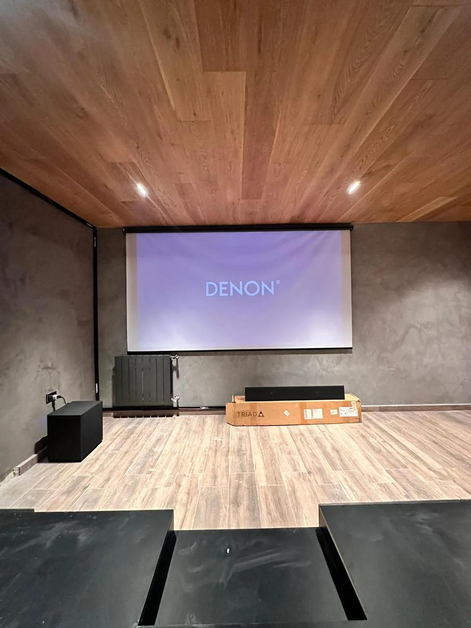Home cinema en obra: pantalla de proyección con el logo de Denon, procesador y barra de parlantes sobre el piso, y la tarima escalonada en primer plano
