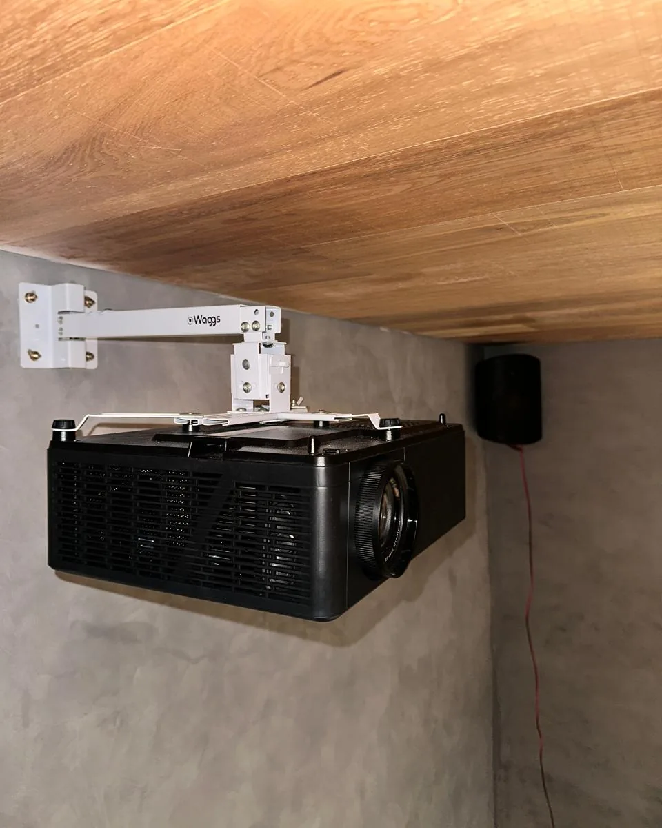 Proyector montado en un soporte a cielorraso de madera, con el parlante surround de pared al lado, en un home cinema en obra