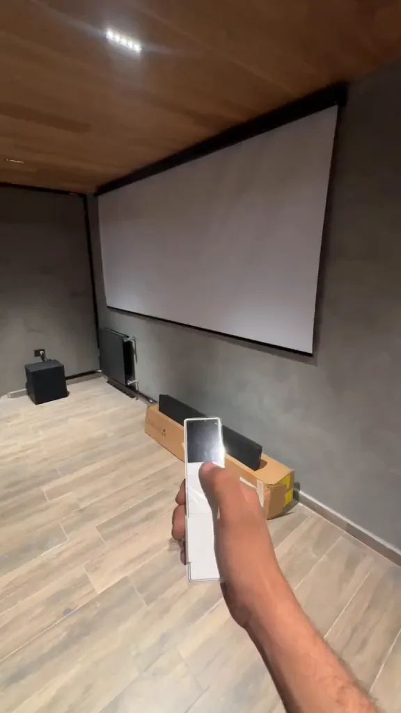 Mano sosteniendo el celular con la app abierta frente a la pantalla de proyección del home cinema, con la barra de parlantes y los surround a la vista